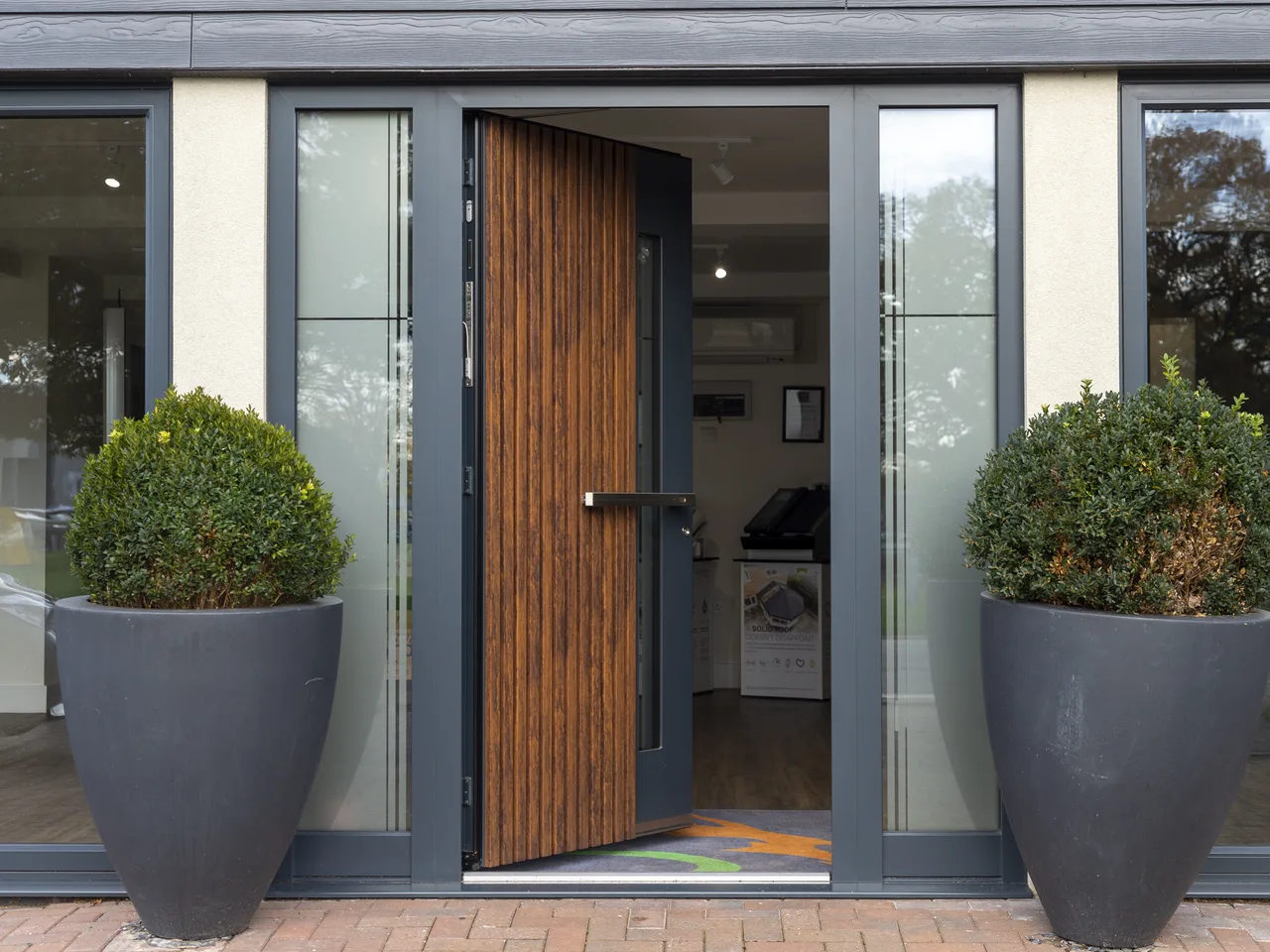 Grey aluminium entrance door with a timber-slat panel flanked by planters
