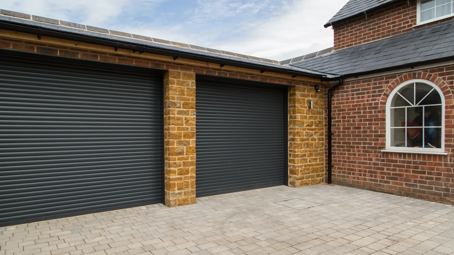 Two anthracite grey roller garage doors between stone piers on a red brick garage with a block-paved drive