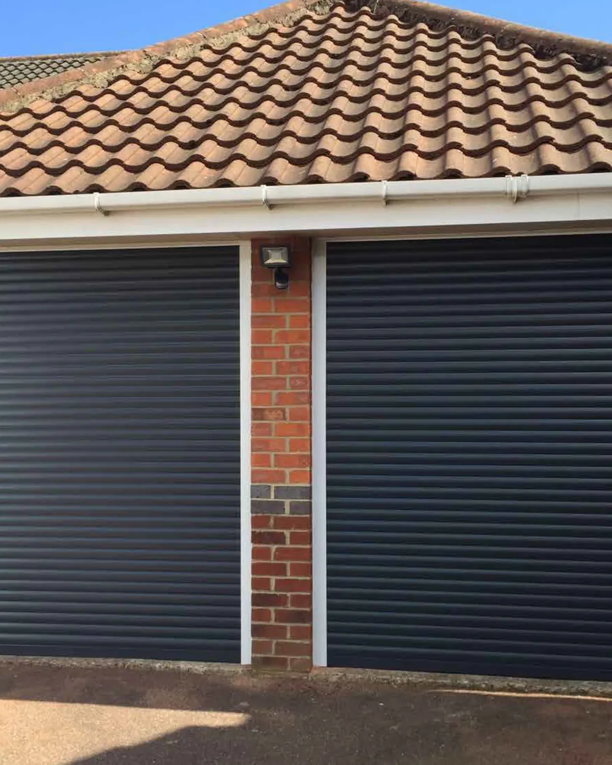 Anthracite grey roller garage door with white guides beside a brick pier on a garage with a tiled roof
