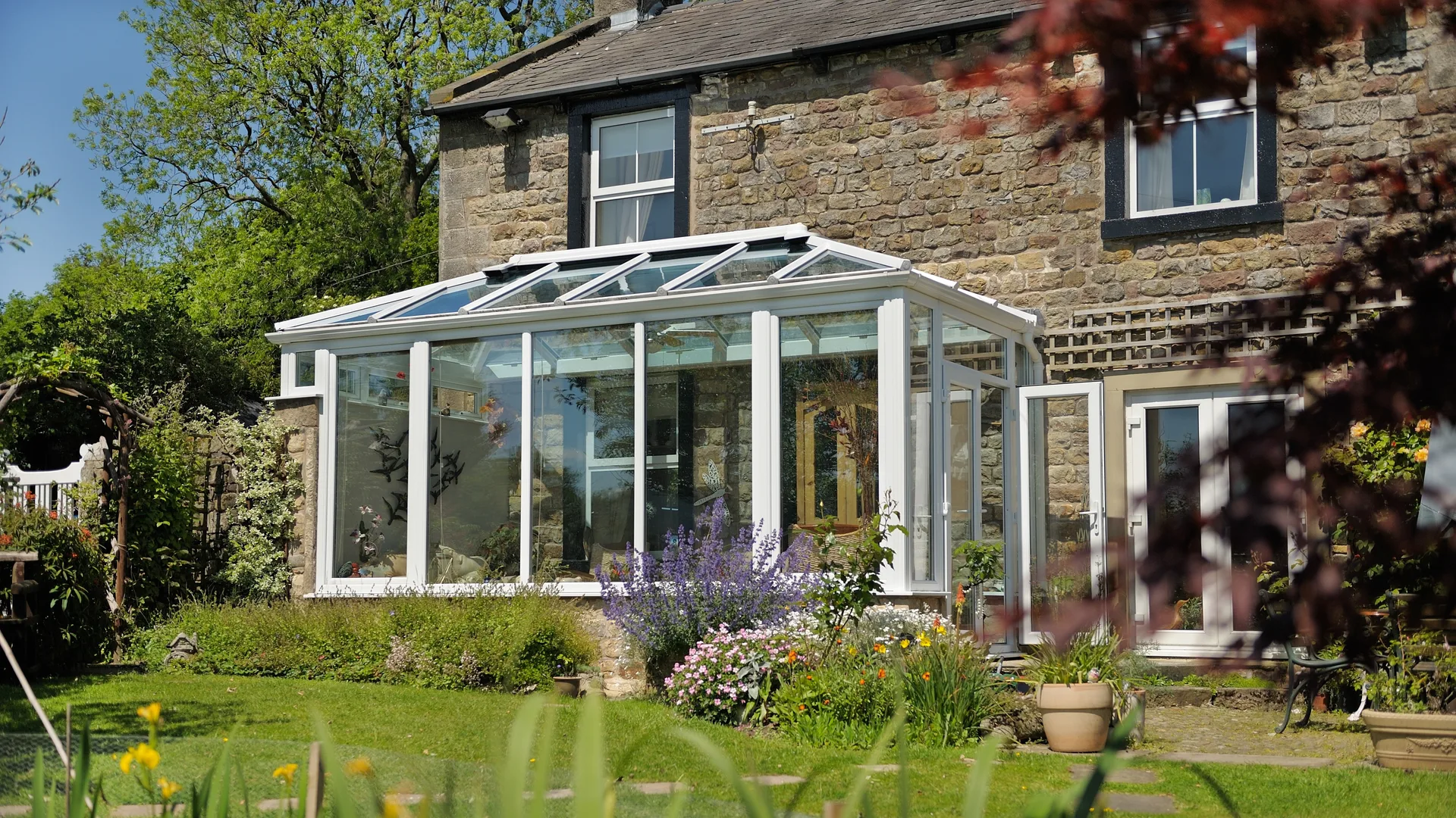 White conservatory with a glazed roof and an open French door on a stone cottage, behind a sunny flower garden