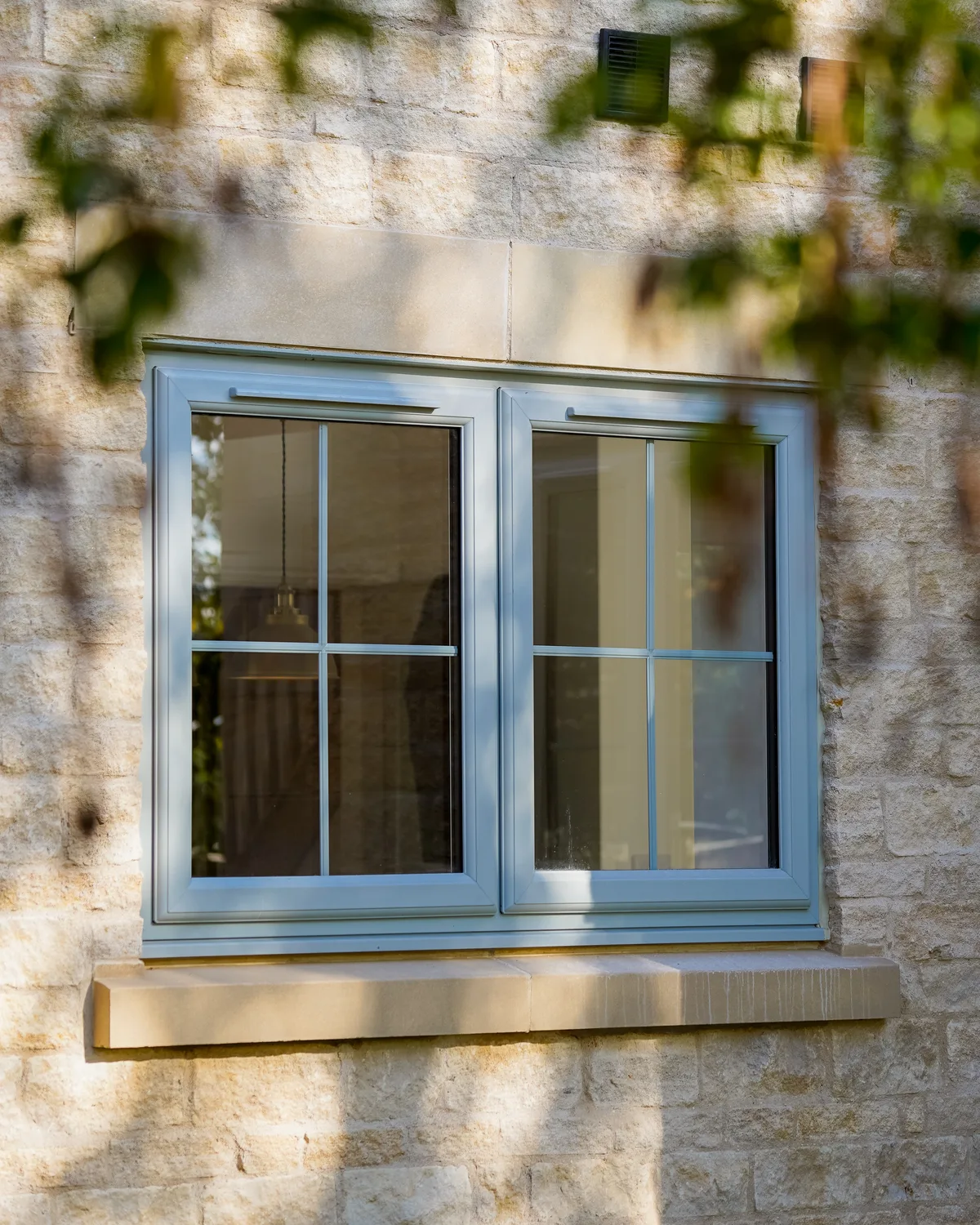 Light grey casement window with Georgian bars in a honey stone wall, framed by soft-focus leaves