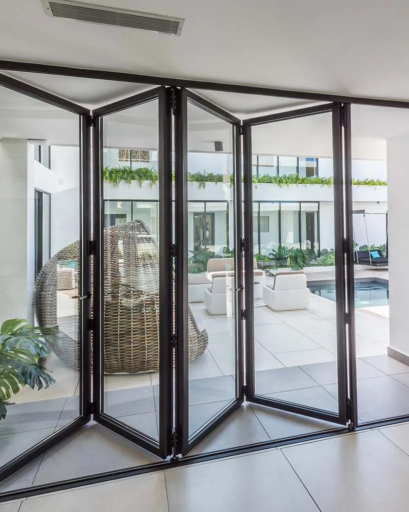 Black aluminium bifold doors part-folded in a bright white room, opening onto a courtyard with a pool