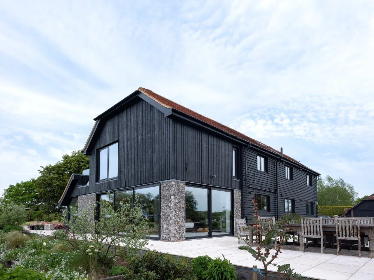 Black timber-clad barn house with black aluminium tilt and turn windows and glazed doors onto a stone terrace