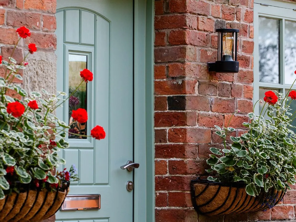 Sage green cottage-style composite door with a small glazed panel beside baskets of red geraniums