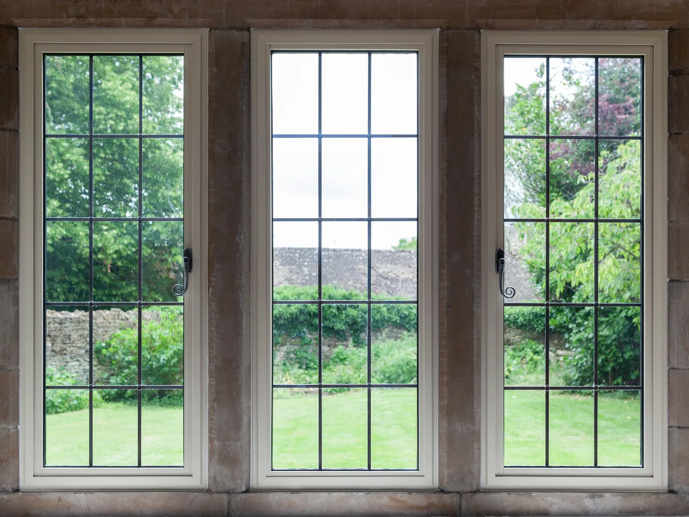 Three cream aluminium heritage windows with slim grid glazing bars between stone mullions, overlooking a garden