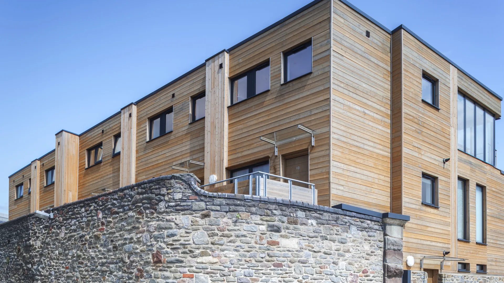 Timber-clad terrace homes with dark grey aluminium tilt and turn windows behind a curved stone wall