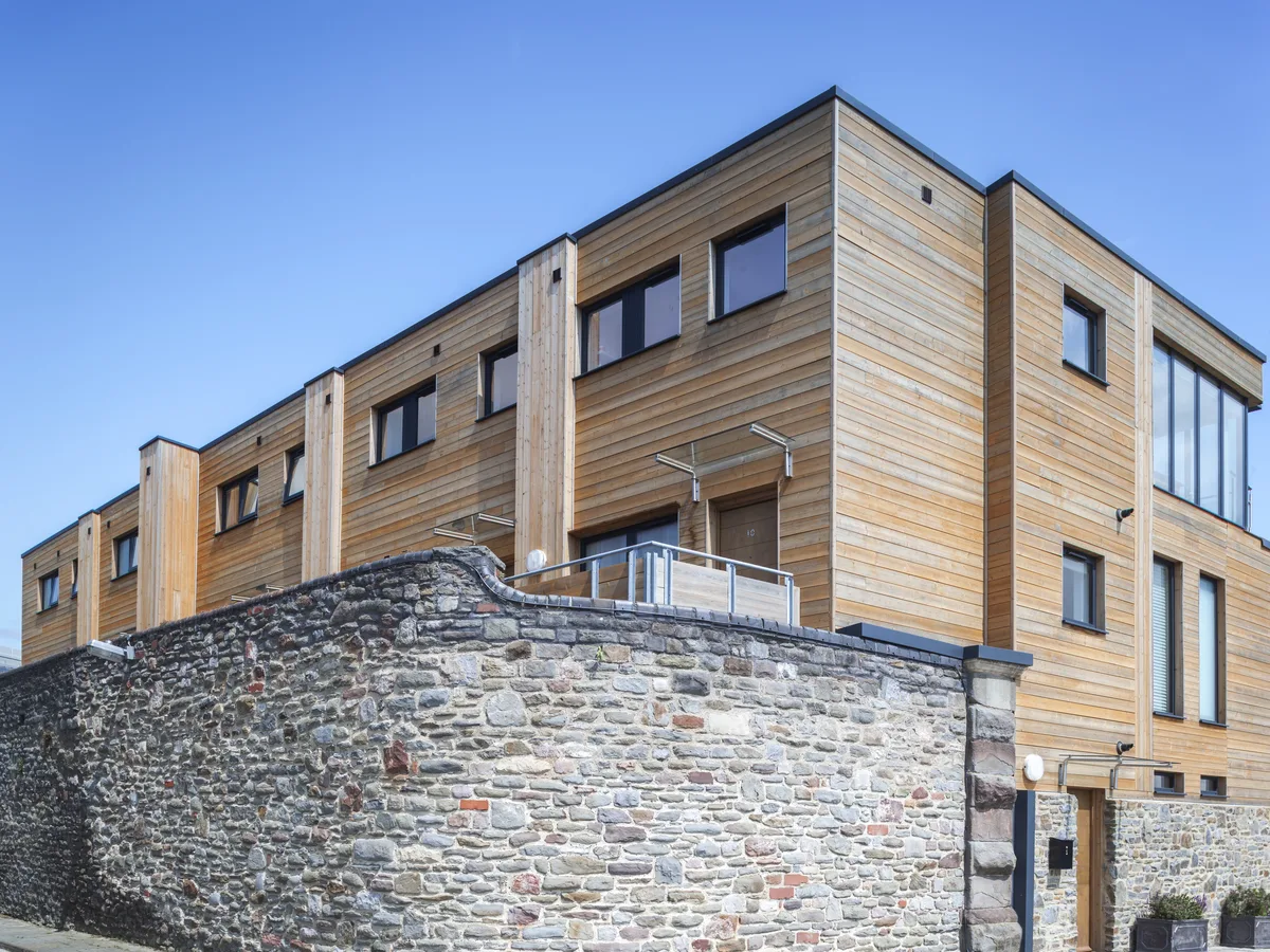 Timber-clad terrace homes with dark grey aluminium tilt and turn windows behind a curved stone wall