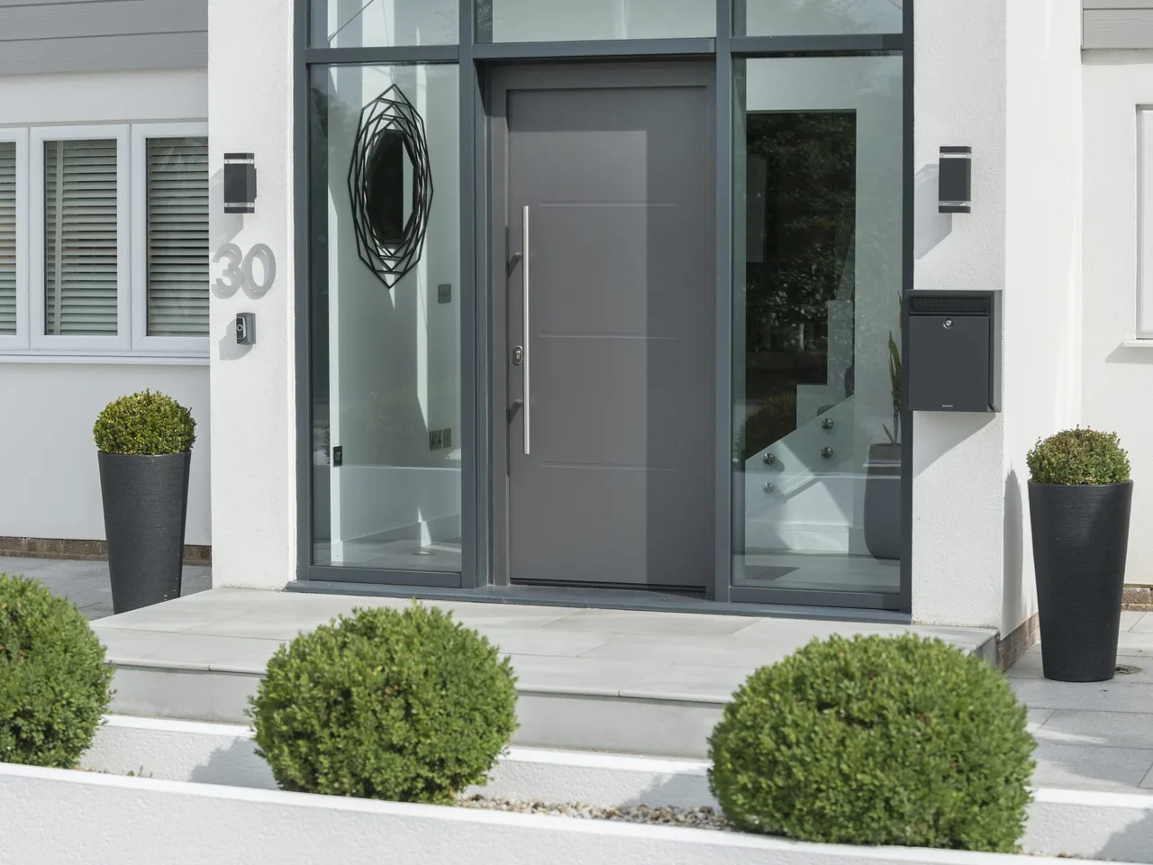 Grey aluminium front door with porthole on a white rendered house, number 30, with planters